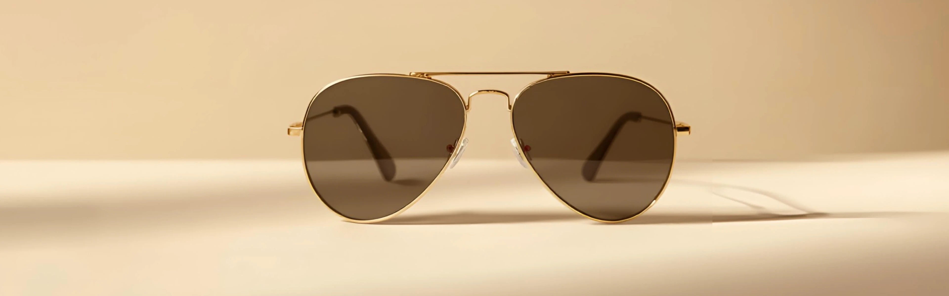 Occhiali da sole modello aviatore con montatura dorata e lenti scure su sfondo beige.